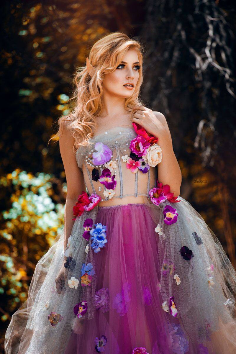 A woman wearing a strapless fantasy gown with a sheer gray tulle bodice, purple tulle skirt, and colorful 3D flowers, standing in a dark forest setting.
