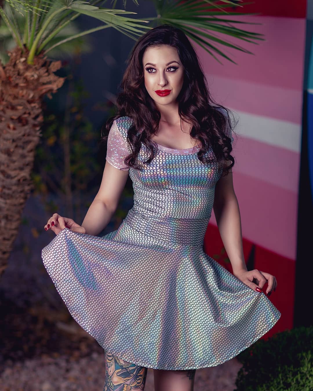 A woman wearing a short-sleeve iridescent holographic skater dress with a textured pattern, posing outdoors with palm leaves and a pink striped background.