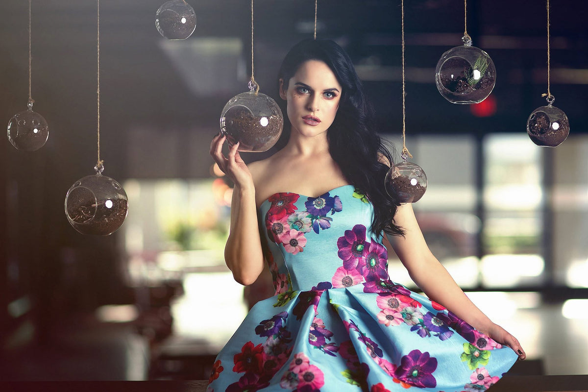 A woman wearing a strapless light blue dress with a vibrant floral print in pink, purple, and green, holding a hanging glass orb in a dimly lit indoor setting.