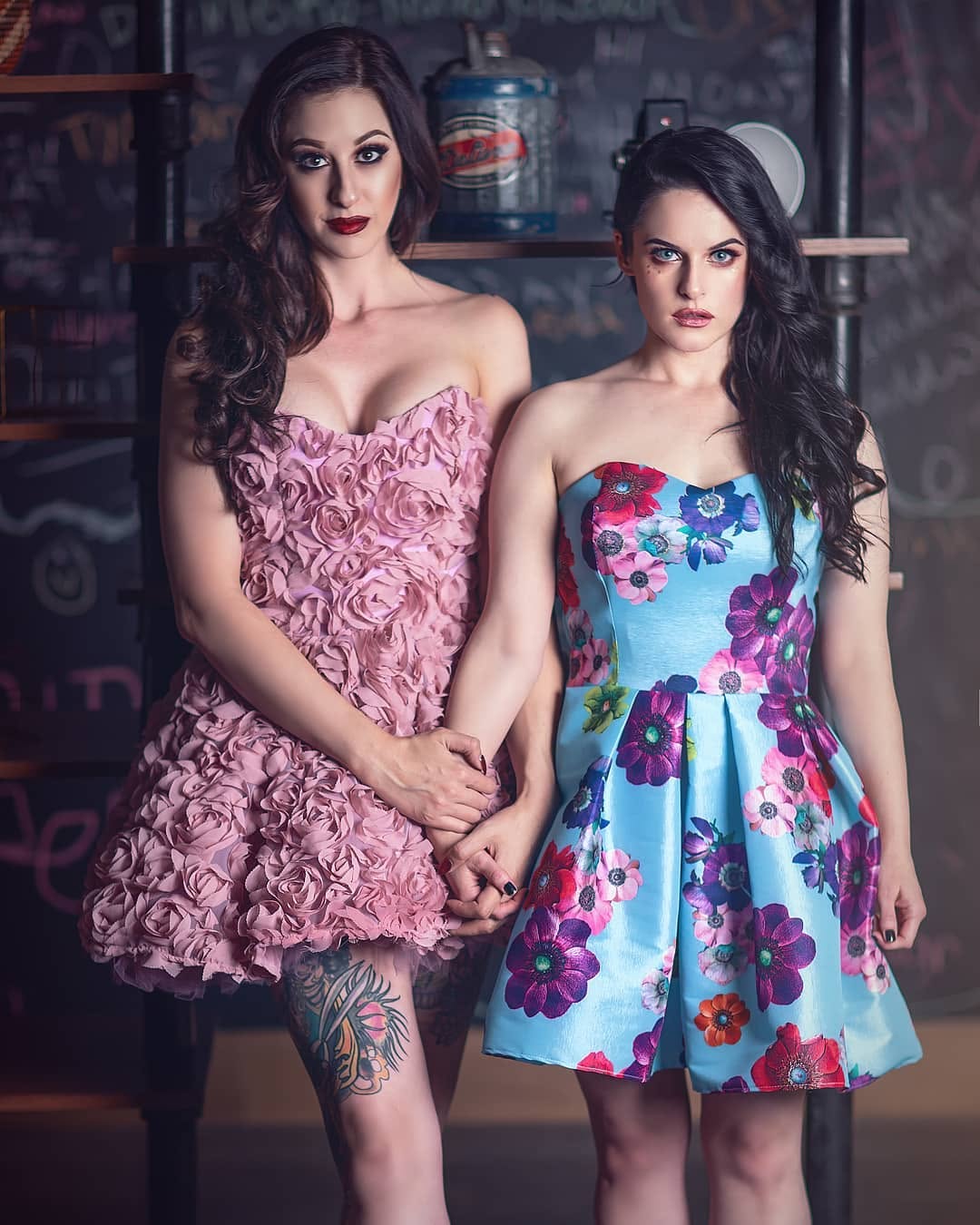 Two women standing side by side holding hands, wearing strapless mini dresses. The woman on the left wears a pink ruffled strapless dress, and the woman on the right wears a light blue strapless dress with a colorful floral print.