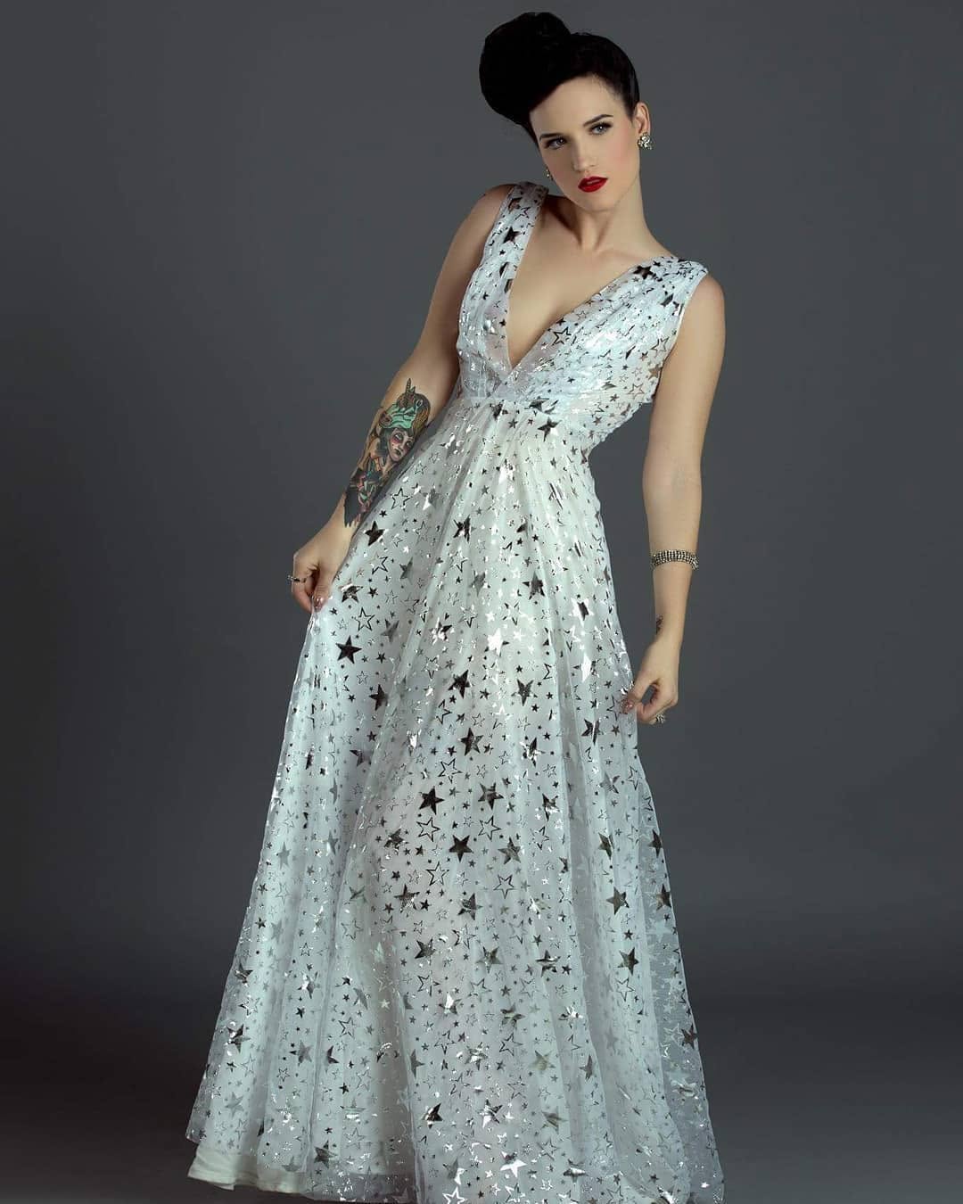 A sleeveless, floor-length gown with a deep V-neckline, featuring an allover pattern of black stars on a shimmering silver base. The model is posed against a dark gray studio background.