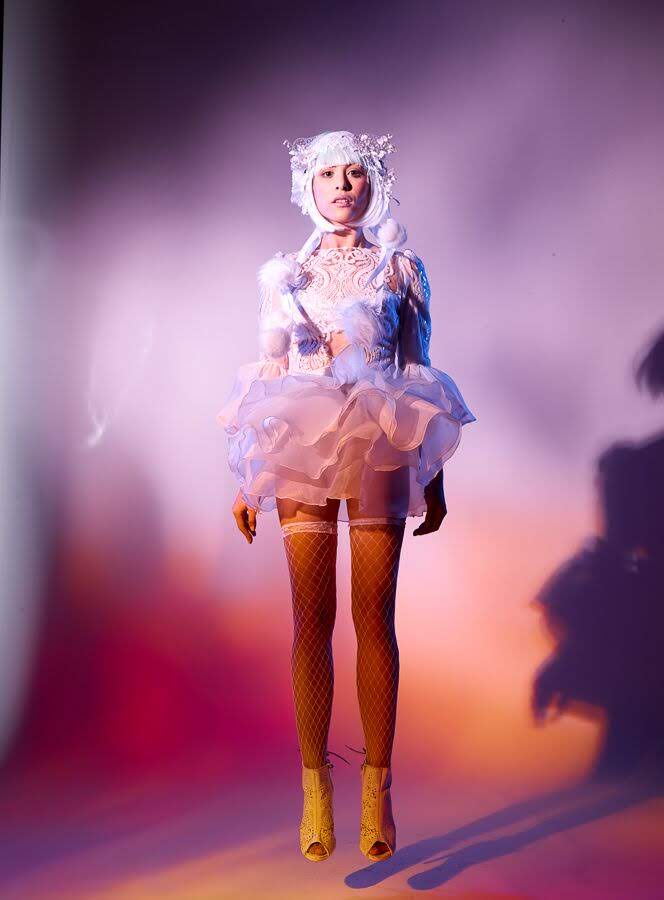 A model stands in a studio wearing a whimsical white and pale pink costume dress with a ruffled tulle skirt, a matching white headpiece, fishnet stockings, and yellow high heels against a purple and pink lit background.