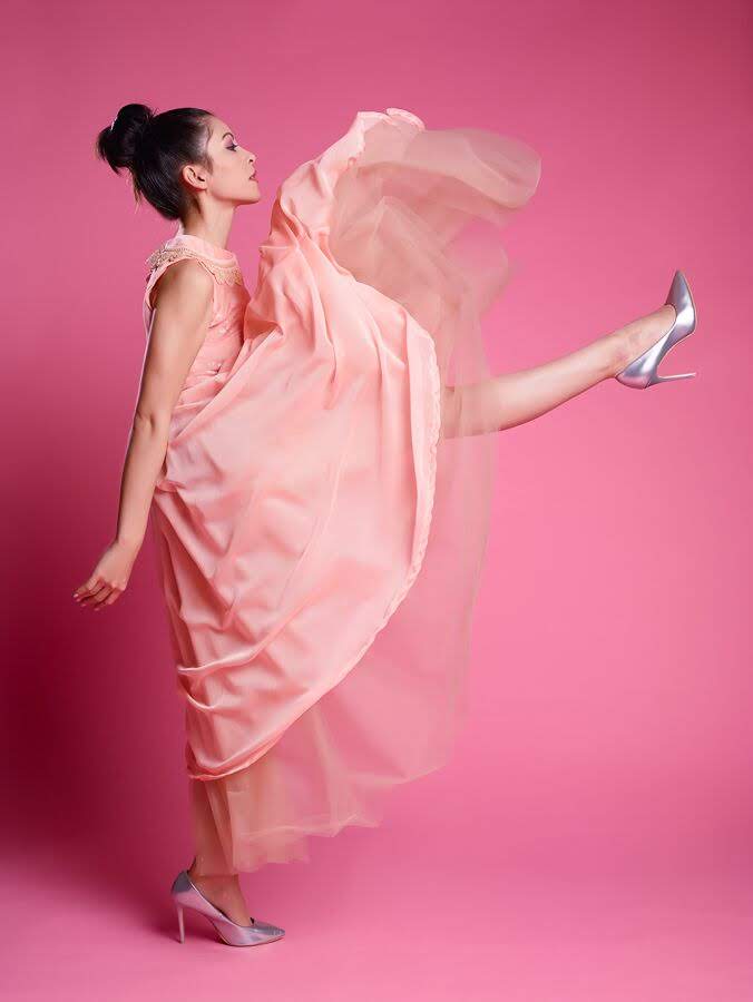 A woman in a flowing light pink gown and silver high heels poses dynamically against a solid pink studio background, with her dress billowing in the wind.
