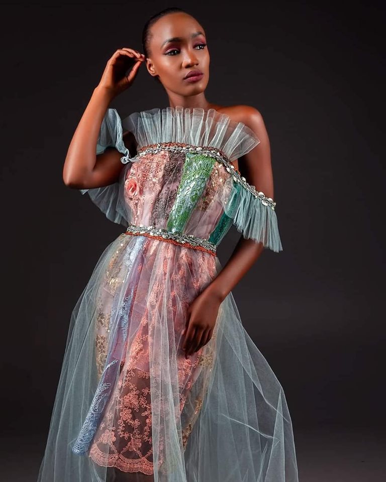 A model wears a highly detailed, multi-layered off-the-shoulder dress featuring sheer tulle, lace, and metallic embellishments in shades of pink, green, and silver, set against a dark studio background.