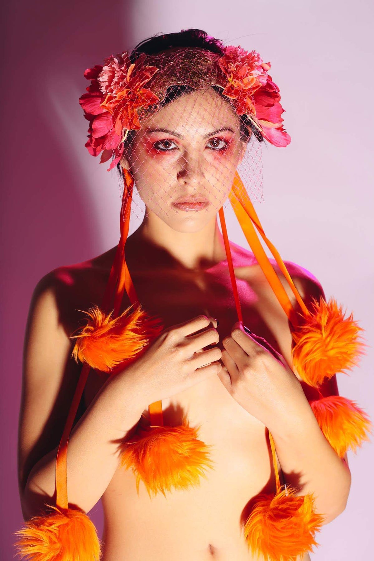 A model wearing a vibrant pink and orange floral headpiece with a netted veil, orange feather boas, and orange eye shadow, posing against a pink studio background.
