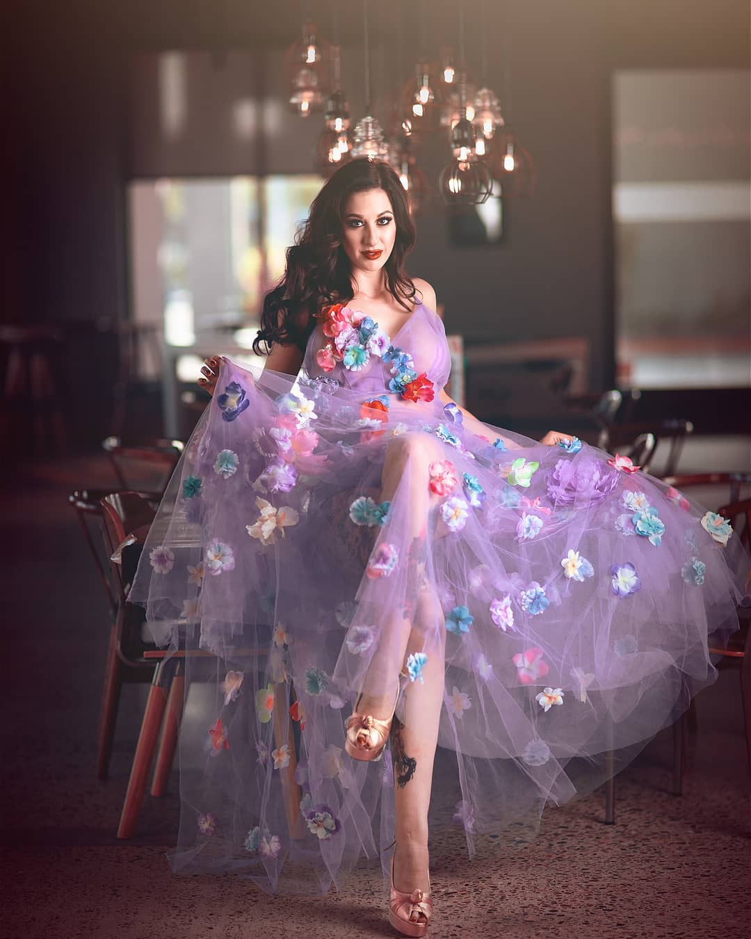 A woman wearing a lavender tulle evening gown with colorful 3D floral appliqués, seated in a dimly lit indoor venue with hanging lights in the background.
